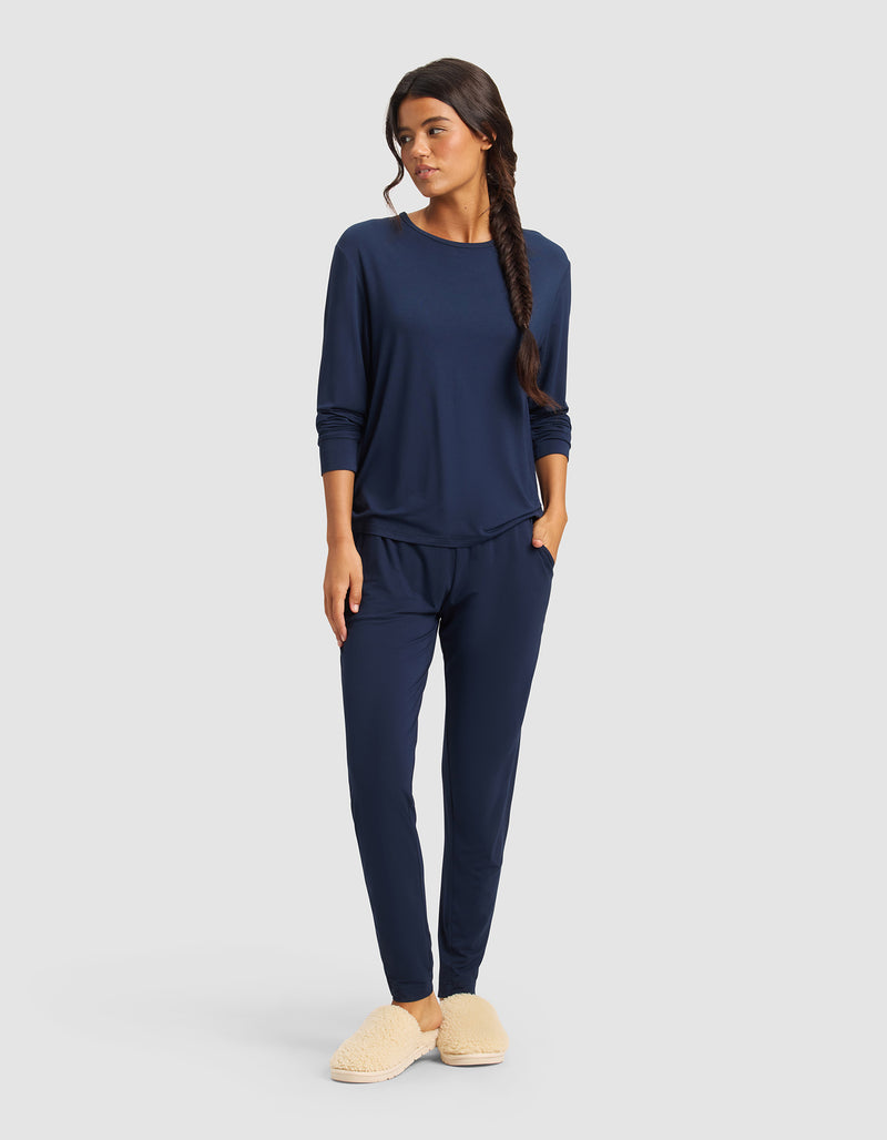 A woman stands with her hands in her pockets, wearing Cozy Earth’s Women's Bamboo Stretch-Knit Tapered Pant in navy blue and beige slippers, looking to the side against a plain light gray background.