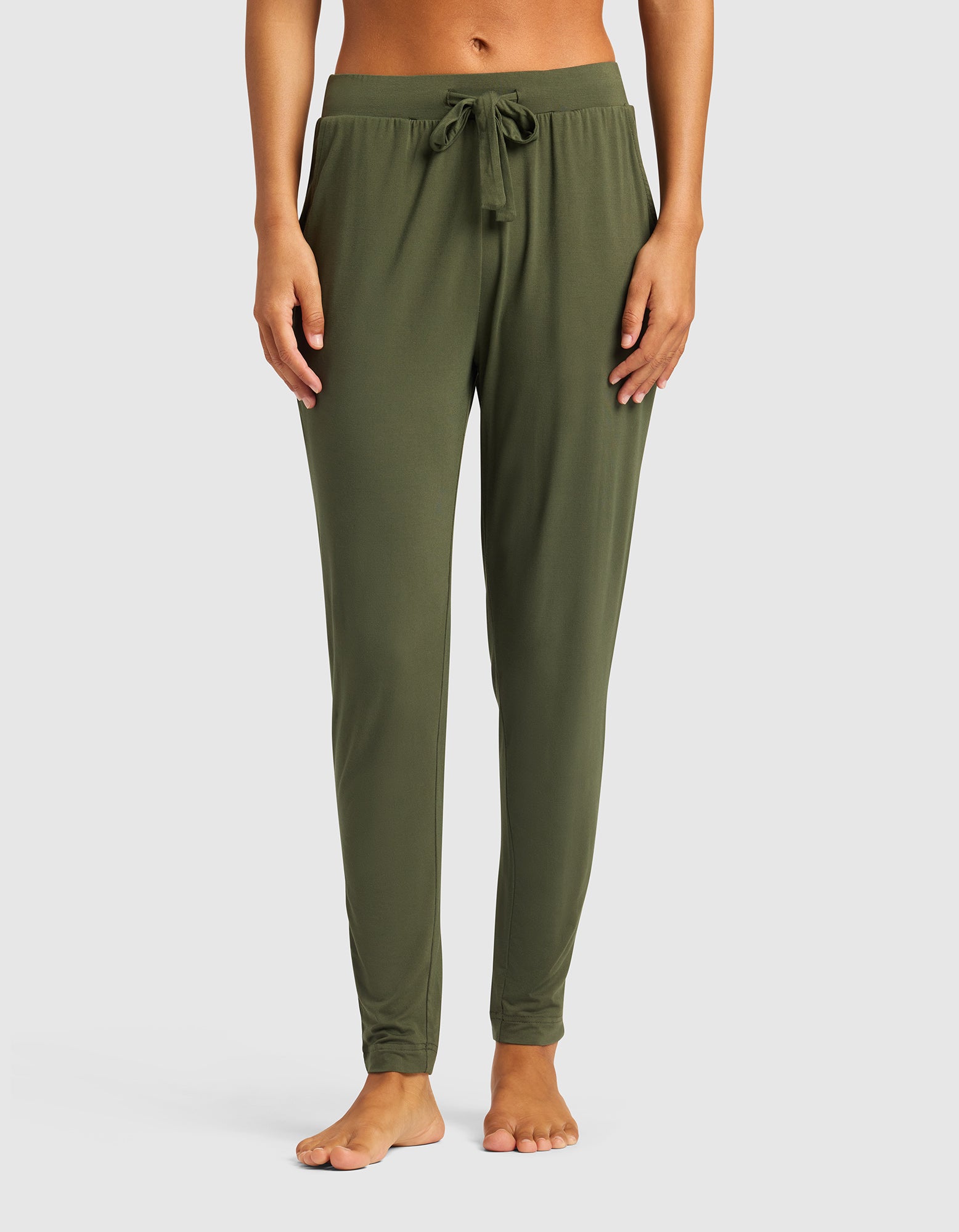 A person stands barefoot against a plain white background, wearing Cozy Earth's Women's Bamboo Stretch-Knit Tapered Pant in olive green with a drawstring waist and bare midriff, shown from shoulders to feet.