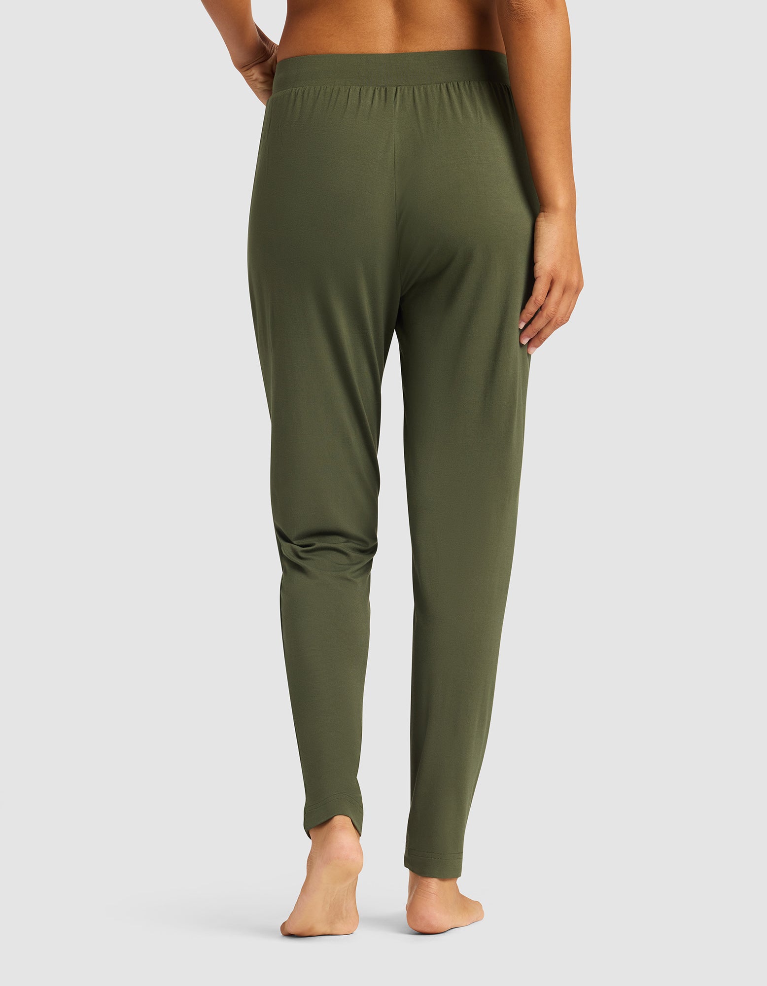 A woman stands barefoot on a white background, shown from the back with one hand on her hip, wearing Cozy Earth's olive green Women's Bamboo Stretch-Knit Tapered Pant.