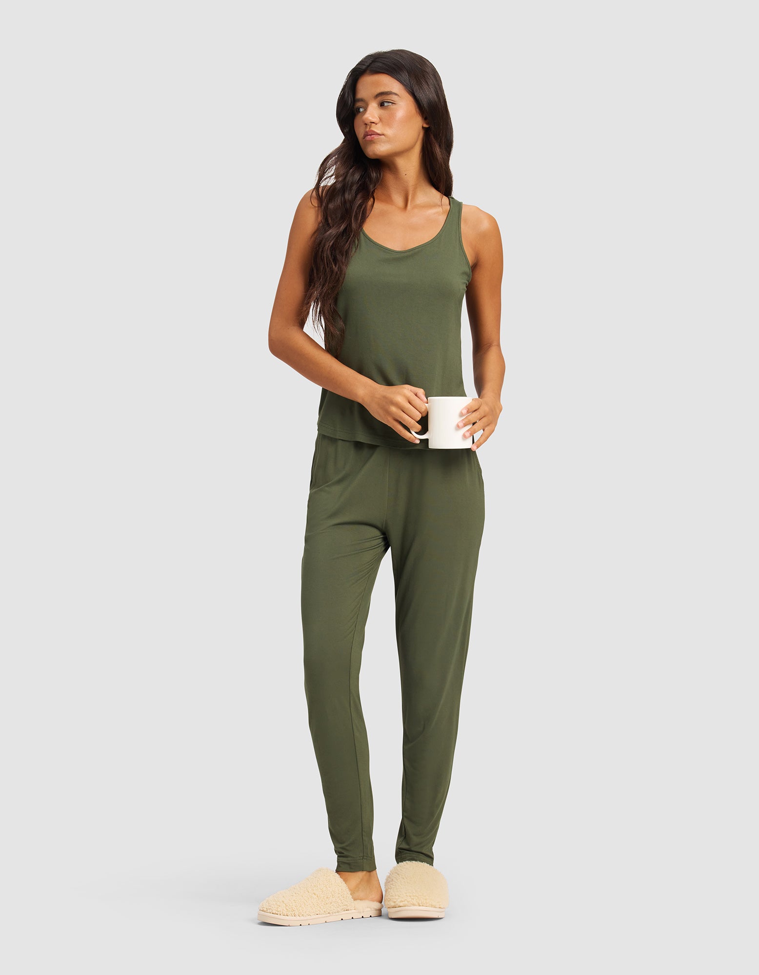 A woman with long dark hair models Cozy Earth's Women's Bamboo Stretch-Knit Tapered Pant in olive green, paired with beige slippers and holding a white mug, standing against a plain light gray background.