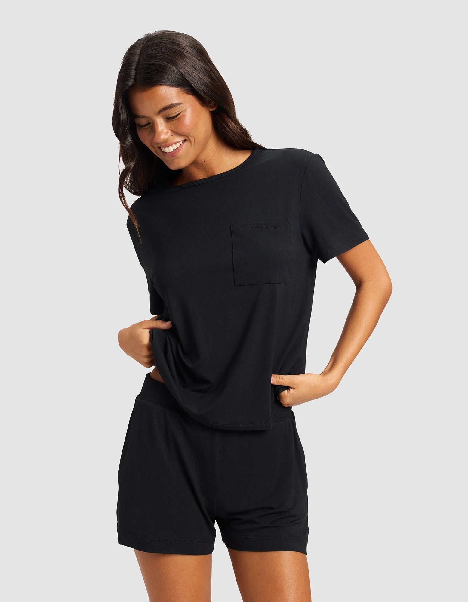 A woman with long dark hair wears the Cozy Earth Women's Bamboo Stretch-Knit Lounge Tee in black, paired with matching shorts. She smiles and looks down, hands in her pockets, standing against a light gray background.