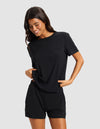 A woman with long dark hair wears the Cozy Earth Women's Bamboo Stretch-Knit Lounge Tee in black, paired with matching shorts. She smiles and looks down, hands in her pockets, standing against a light gray background.