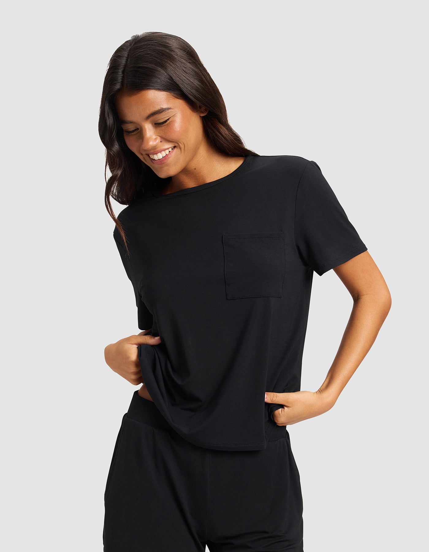 A woman with long dark hair smiles and looks down while wearing the Cozy Earth Women's Bamboo Stretch-Knit Lounge Tee & Pant Set in black against a plain light gray background.