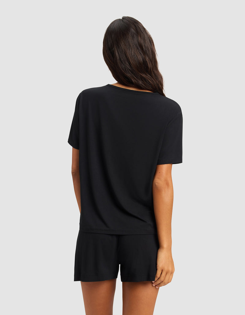 A woman with long brown hair stands with her back to the camera, wearing the Cozy Earth Women's Bamboo Stretch-Knit Lounge Tee in black, paired with matching shorts, against a plain light gray background.