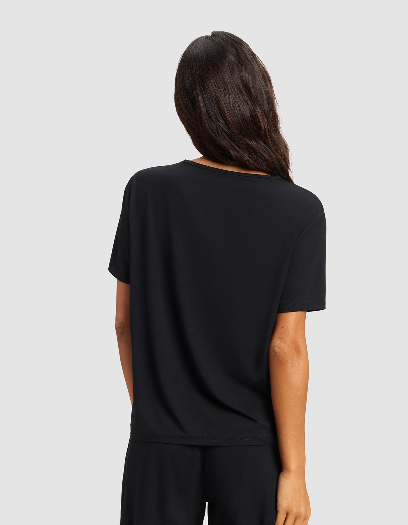 A woman with long, dark hair stands with her back to the camera, wearing Cozy Earth’s Women's Bamboo Stretch-Knit Lounge Tee & Pant Set in black against a light gray background.