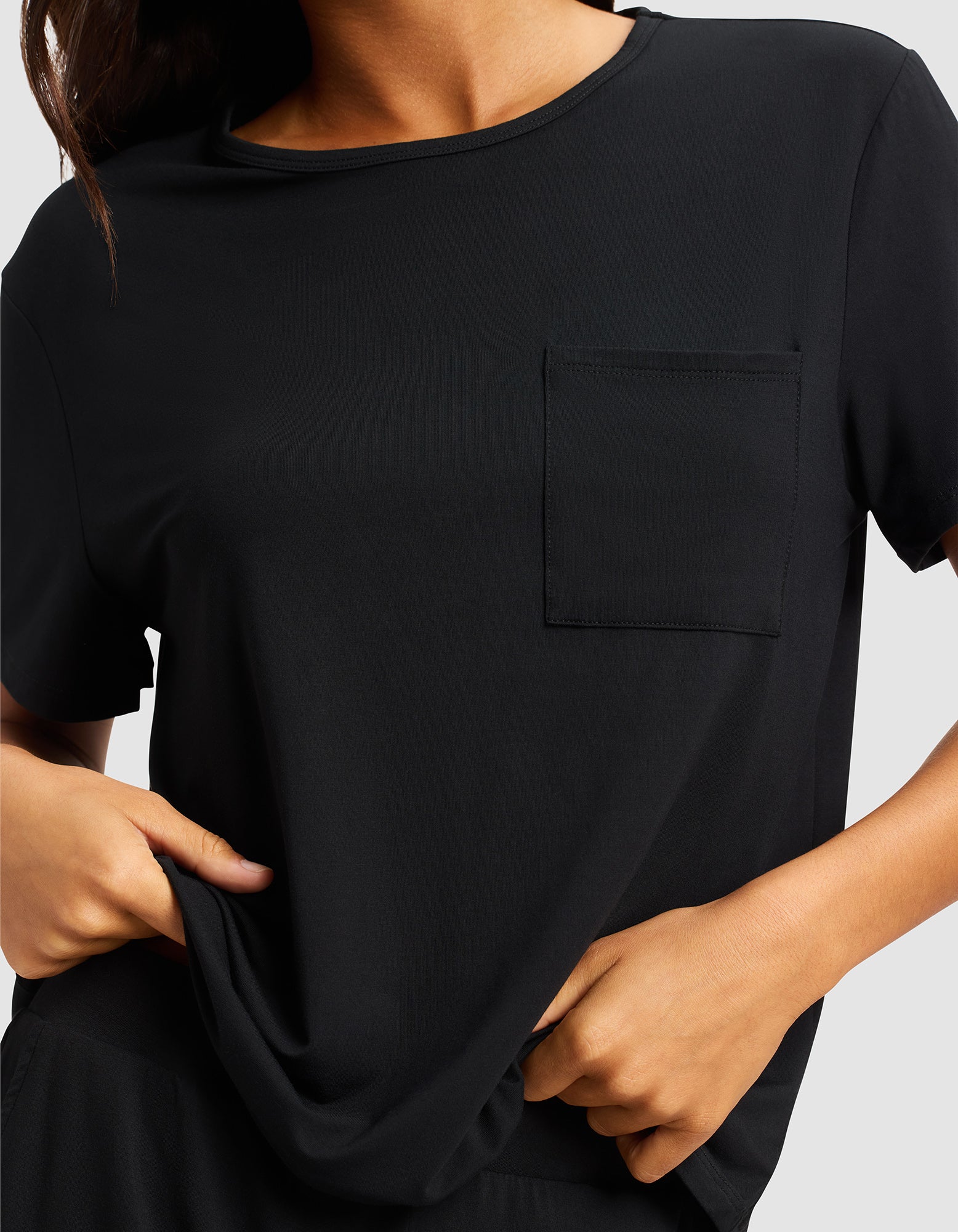 A woman is shown from shoulders to waist wearing the Cozy Earth Women's Bamboo Stretch-Knit Lounge Tee, a plain black short-sleeve shirt with a chest pocket, gently pulling the hem with both hands.