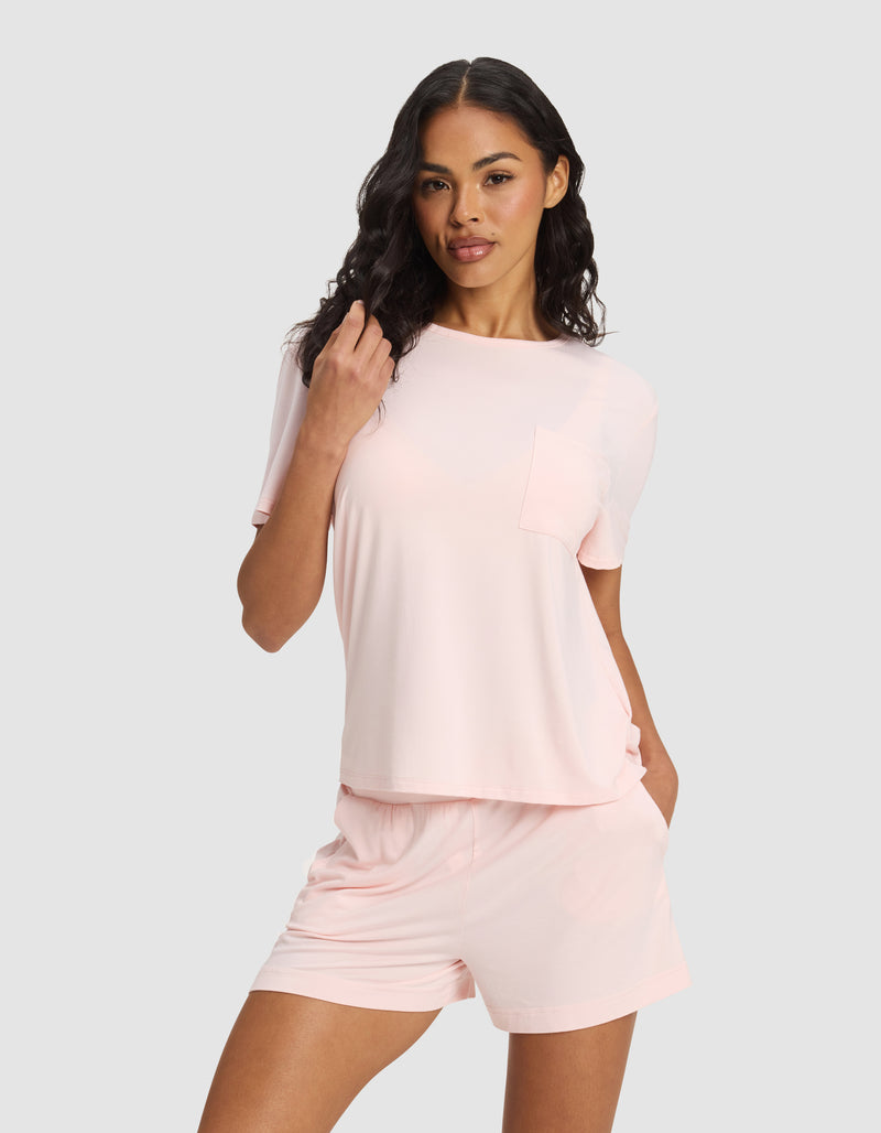 A woman with long, wavy dark hair and a relaxed, confident look stands against a plain background wearing Cozy Earth's Women's Bamboo Stretch-Knit Lounge Tee in light pink with a pocket, paired with matching pink shorts.