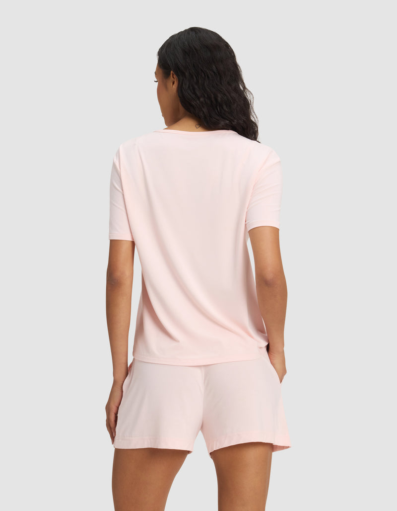 A woman with wavy dark hair is seen from the back, modeling the Cozy Earth Women's Bamboo Stretch-Knit Lounge Tee in light pink, paired with matching shorts against a plain light gray background.