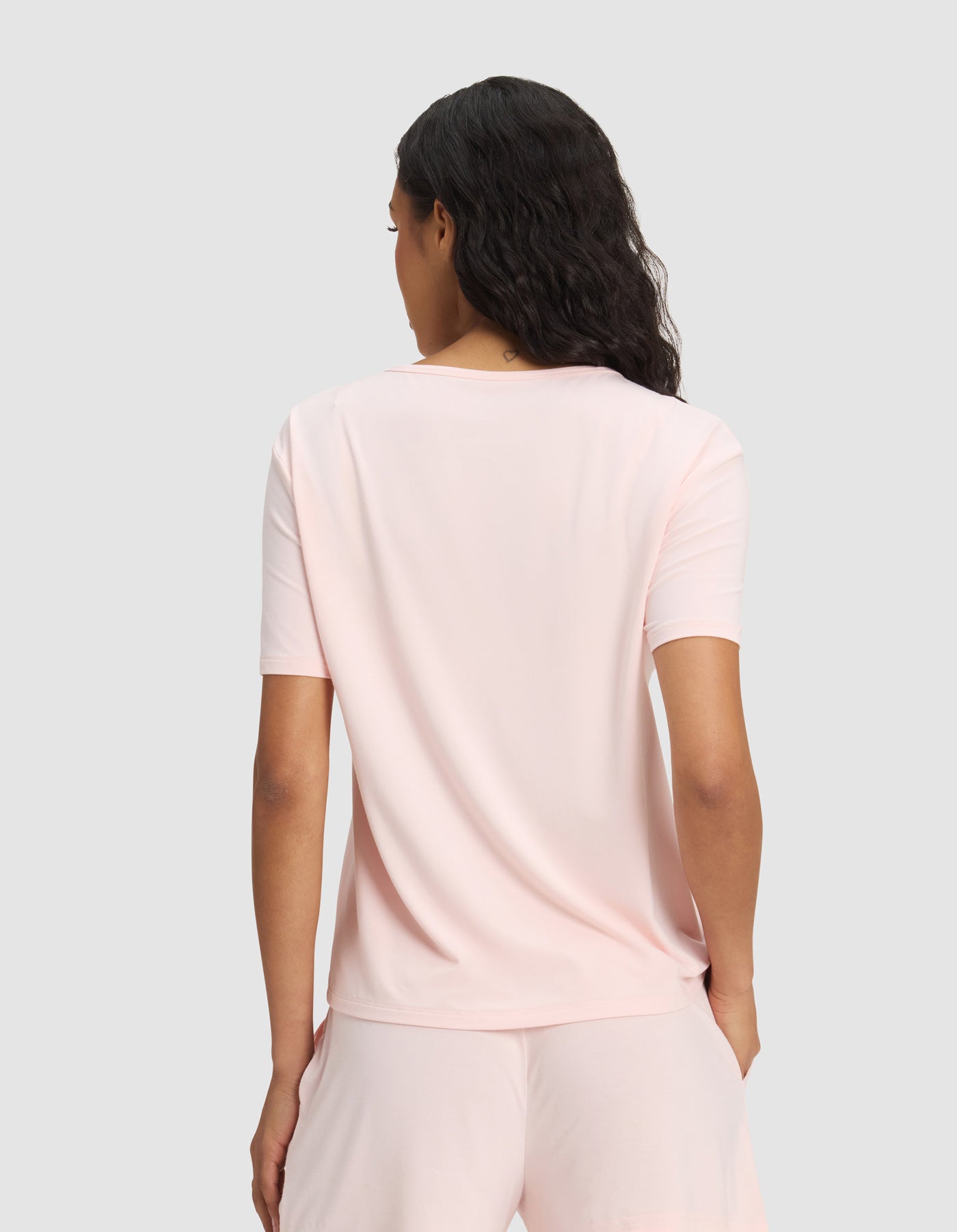 A woman with wavy dark hair is seen from the back wearing the Cozy Earth Women's Bamboo Stretch-Knit Lounge Tee & Pant Set in light pink, against a plain, light gray background.