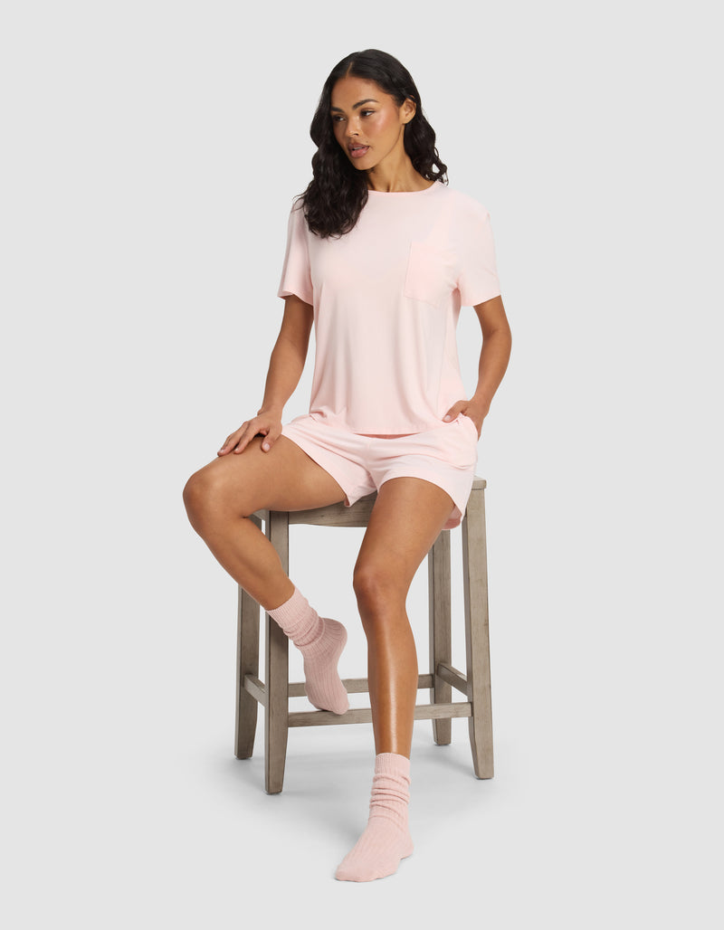 A woman with dark wavy hair sits on a wooden stool, wearing Cozy Earth’s Women's Bamboo Stretch-Knit Lounge Tee in light pink, matching shorts, and pink socks. She looks to the side with one leg on the stool and the other foot on the floor.