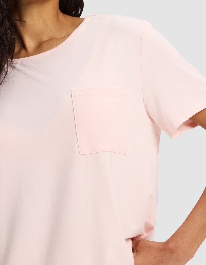 A woman models the Cozy Earth Women's Bamboo Stretch-Knit Lounge Tee in light pink, featuring short sleeves and a chest pocket, posing with one hand on her hip against a plain light background. Only her upper torso is shown.