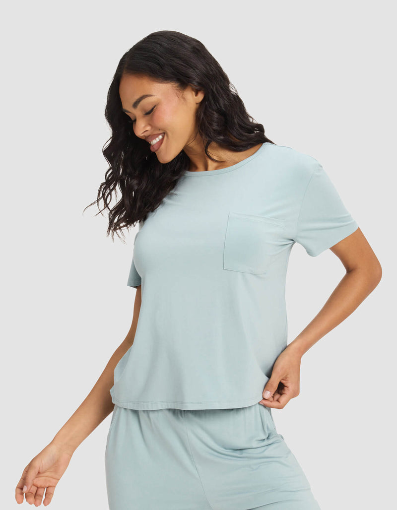 A woman with wavy dark hair smiles and looks down while wearing the Cozy Earth Women’s Bamboo Stretch-Knit Lounge Tee & Short Set, featuring a light blue color, chest pocket, short sleeves, and a relaxed fit.