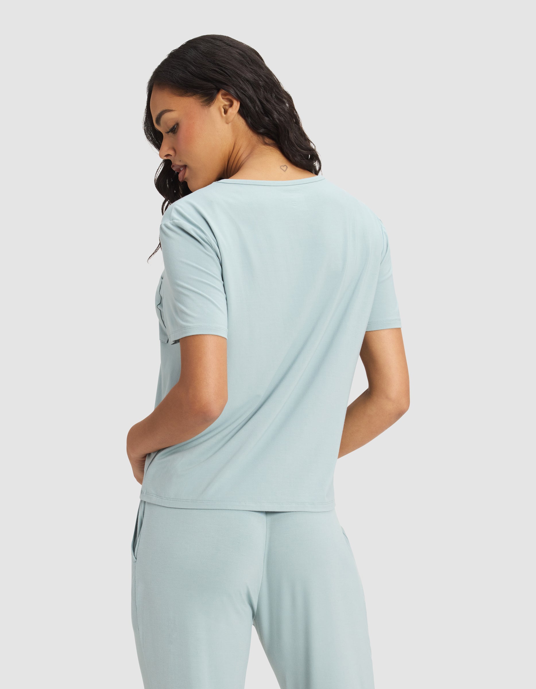 A woman faces away in a Cozy Earth Women's Bamboo Stretch-Knit Lounge Tee, light blue with short sleeves and matching pants. She turns her head slightly, hands in pockets, standing against a plain light background.