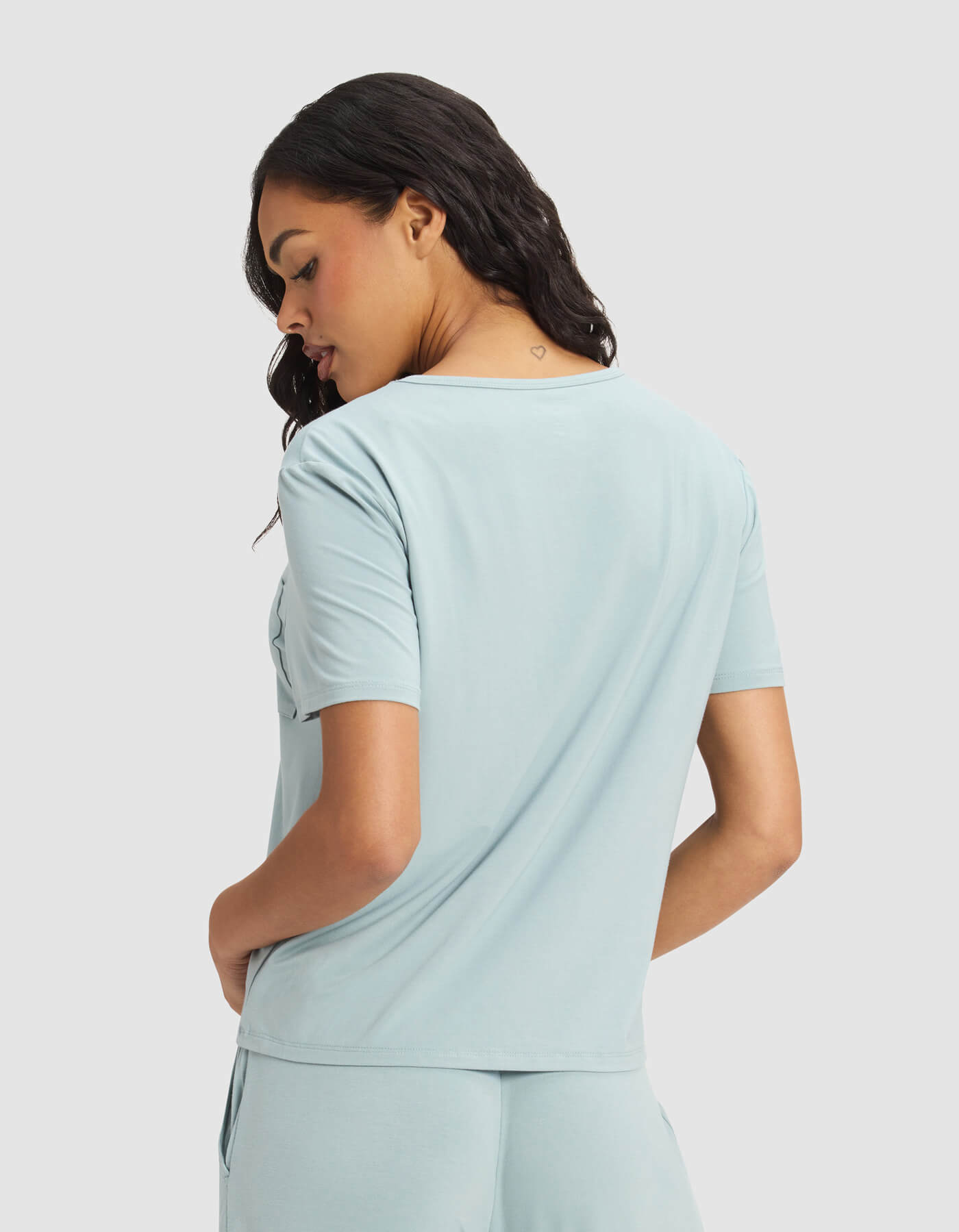 A woman with wavy dark hair is seen from the back, wearing the Cozy Earth Women's Bamboo Stretch-Knit Lounge Tee & Short Set in light blue, looking down over her shoulder against a plain background.