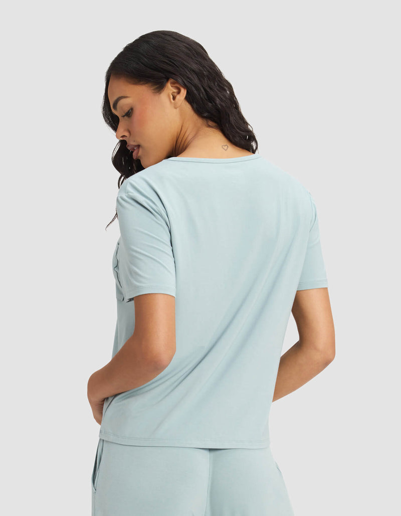 A woman with wavy dark hair is seen from the back, wearing the Cozy Earth Women's Bamboo Stretch-Knit Lounge Tee & Short Set in light blue, looking down over her shoulder against a plain background.