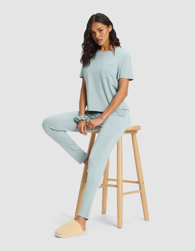 A woman wears the Cozy Earth Women's Bamboo Stretch-Knit Lounge Tee in light blue and cream slippers, sitting on a wooden stool with one foot down and the other leg bent, posing against a plain light background.
