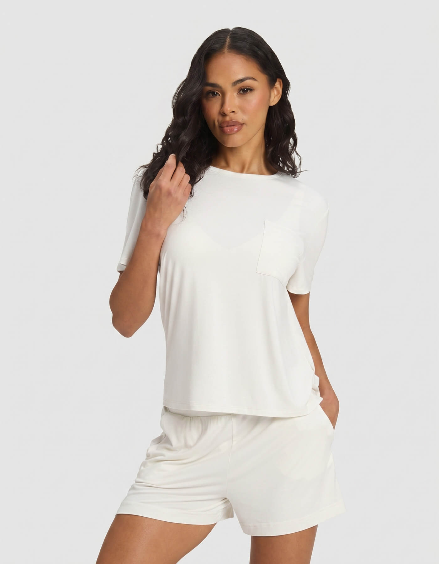 A woman wearing the Cozy Earth Women's Bamboo Stretch-Knit Lounge Tee in Ivory stands against a plain light background, pairing the top with matching shorts, one hand in her pocket and the other touching her hair.