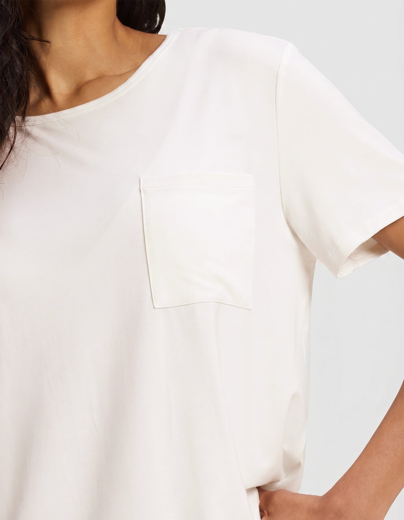 A woman models the Cozy Earth Women's Bamboo Stretch-Knit Lounge Tee, a plain white short-sleeve top with a chest pocket, shown from shoulders to midsection against a light gray background.