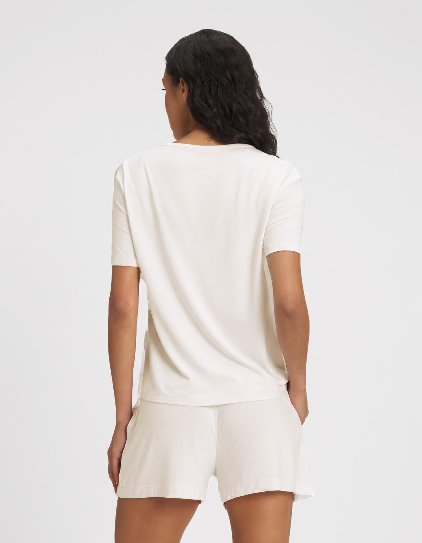 A person with long, wavy dark hair faces away from the camera, wearing Cozy Earth's Women's Bamboo Stretch-Knit Lounge Tee in light beige and matching shorts against a plain white background.