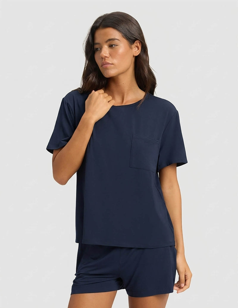 A woman with long dark hair models the Cozy Earth Women's Bamboo Stretch-Knit Lounge Tee & Short Set in navy, featuring a short-sleeve top with chest pocket and matching shorts, against a plain light gray background.
