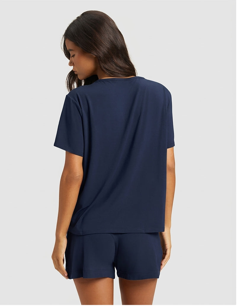 A woman with long dark hair stands facing away in a Cozy Earth Women's Bamboo Stretch-Knit Lounge Tee & Short Set, featuring a loose-fitting navy blue short-sleeve top and matching shorts against a plain light gray background.