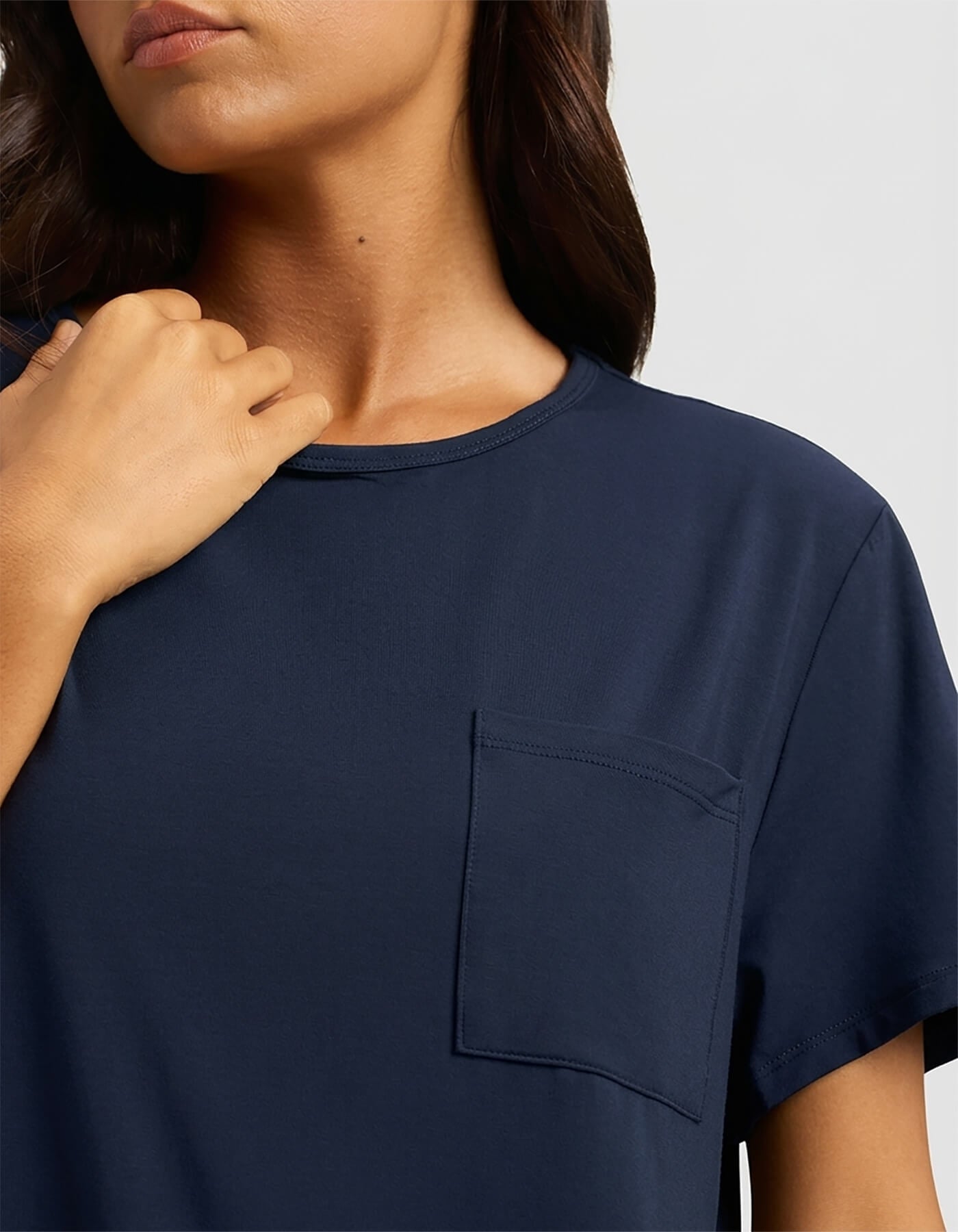 A woman with long brown hair touches her collarbone while wearing the Cozy Earth Women's Bamboo Stretch-Knit Lounge Tee, a navy blue short-sleeve shirt with a chest pocket, shown from shoulders to mid-torso.