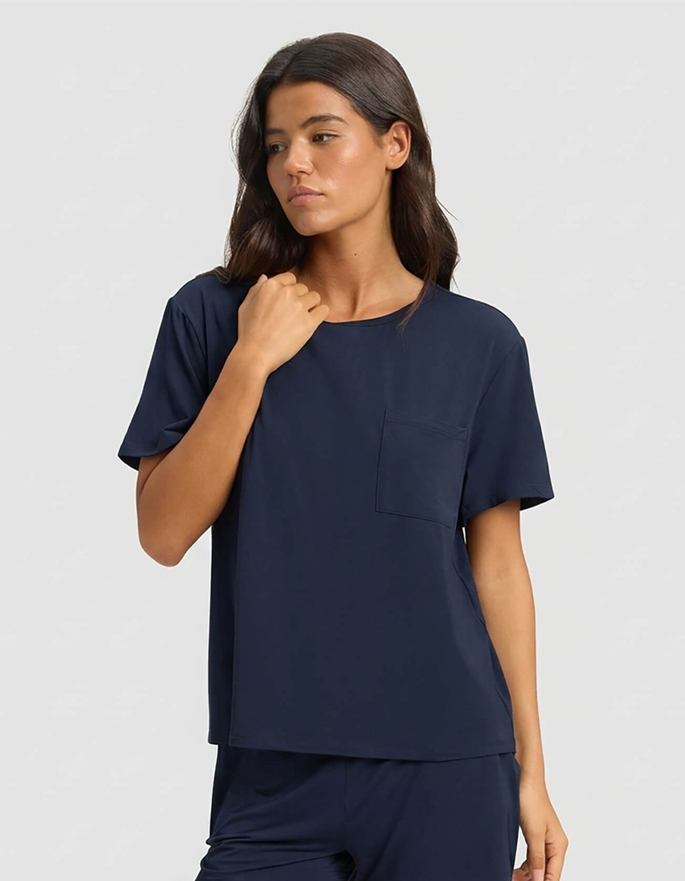 A woman wears the Cozy Earth Women's Bamboo Stretch-Knit Lounge Tee & Pant Set in navy blue, featuring a loose fit and short sleeves with a chest pocket, standing against a light gray background with a neutral expression.|Color:Navy