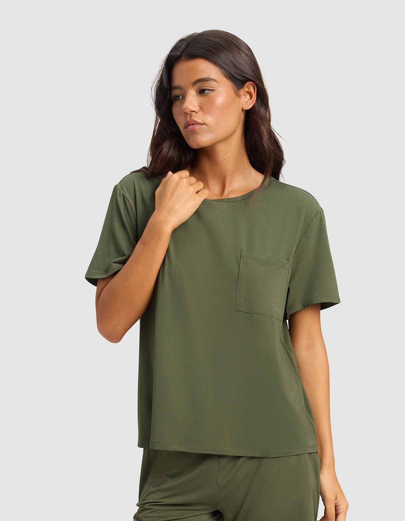 A woman with long dark hair wears the Cozy Earth Women's Bamboo Stretch-Knit Lounge Tee & Pant Set in olive green, featuring a short-sleeve top with chest pocket and matching pants, standing against a plain light gray background.
