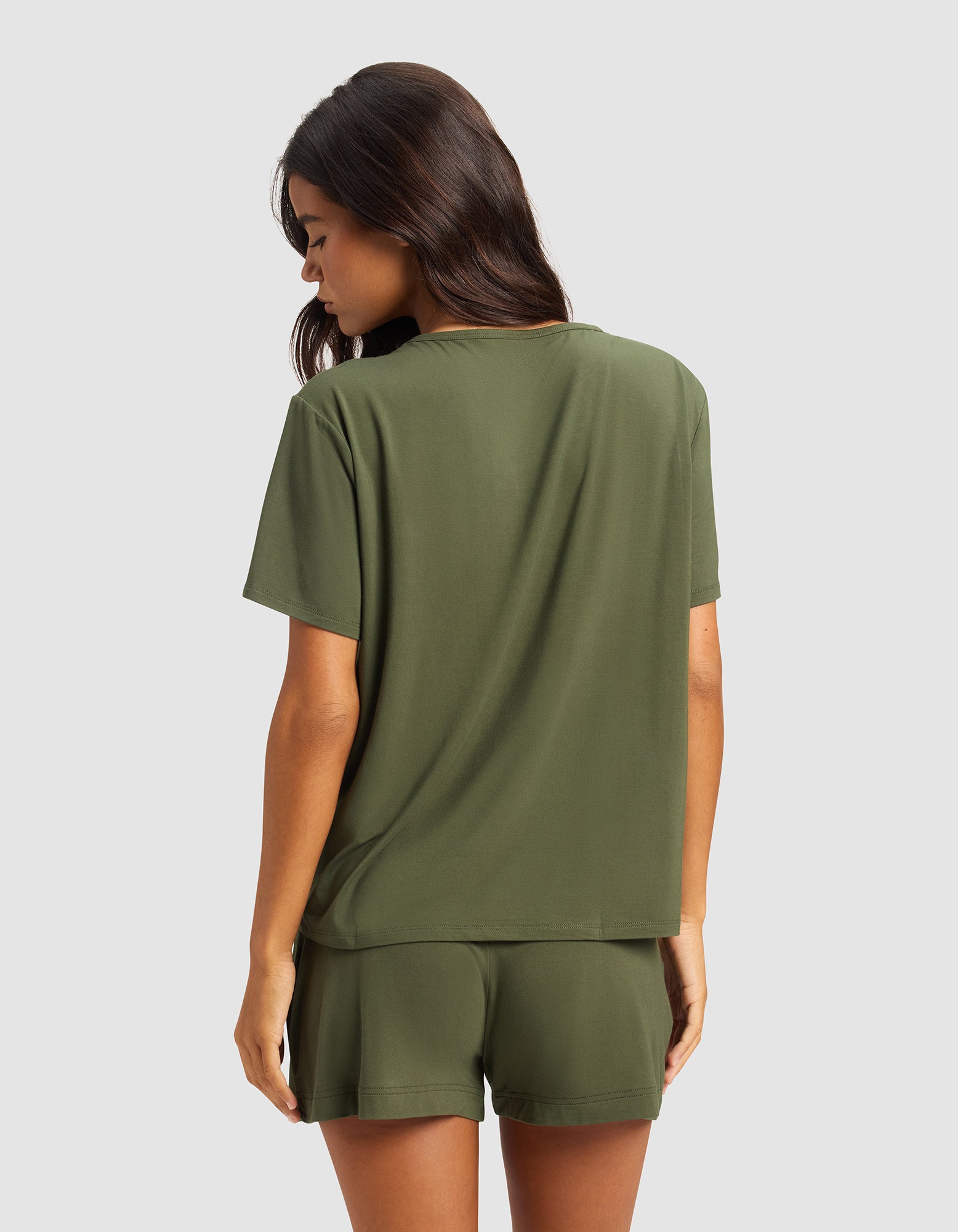 A woman with long brown hair faces away, wearing Cozy Earth's Women's Bamboo Stretch-Knit Lounge Tee in olive green, paired with matching shorts, set against a plain light gray background.