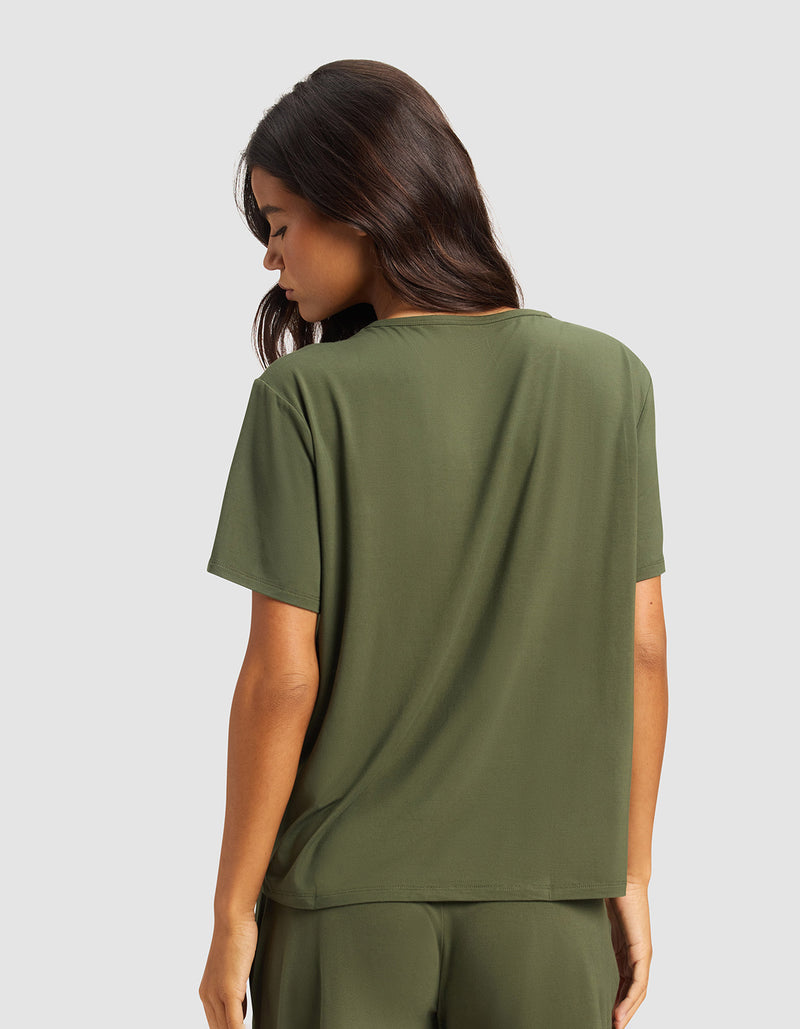 A woman with long dark hair stands with her back to the camera, wearing the Cozy Earth Women's Bamboo Stretch-Knit Lounge Tee & Pant Set in olive green against a plain light gray background.