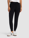 A person stands against a light gray background with hands in pockets, wearing Cozy Earth's Women's Brushed Bamboo Jogger Pant in black, paired with a white shirt and white sneakers.|Color:Black