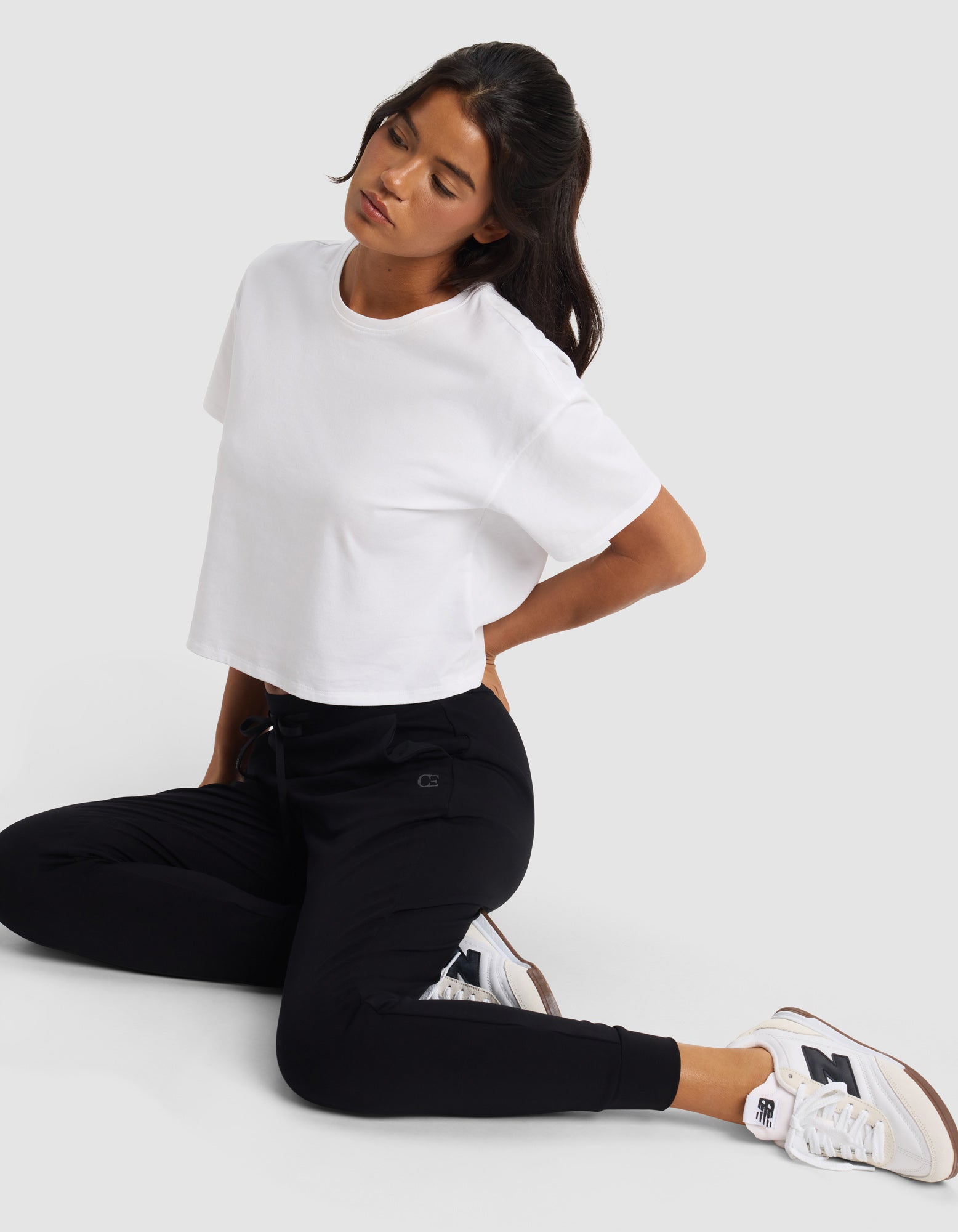 A person wearing a white cropped T-shirt, Cozy Earth Women's Brushed Bamboo Jogger Pant in black, and white New Balance sneakers sits on the floor with one knee up, looking down with a neutral expression against a plain background.