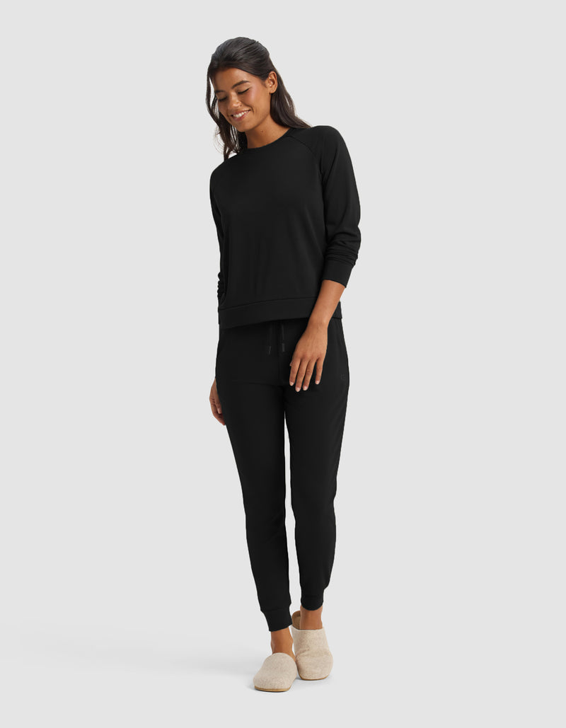A woman smiles, wearing the Cozy Earth Women's Brushed Bamboo Jogger Set in black with jogger pants and beige slippers, standing against a plain light gray background.