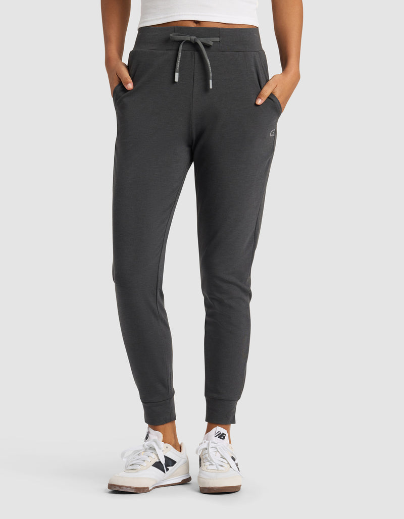 A woman wears Cozy Earth's Women's Brushed Bamboo Jogger Pant in dark gray with white sneakers featuring black accents, standing hands in pockets against a plain light gray background.