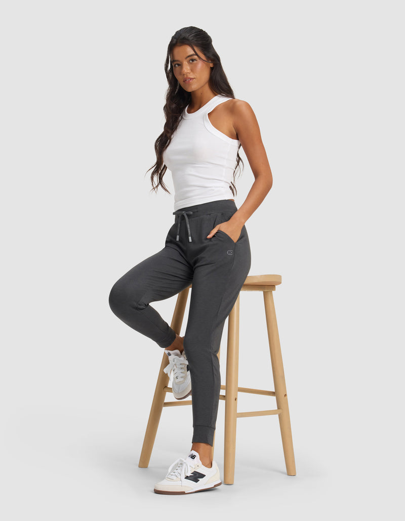 A woman wearing Cozy Earth's Women's Brushed Bamboo Jogger Pant in dark gray poses with one leg on a wooden stool, hands in pockets, and white sneakers, set against a plain light gray background.