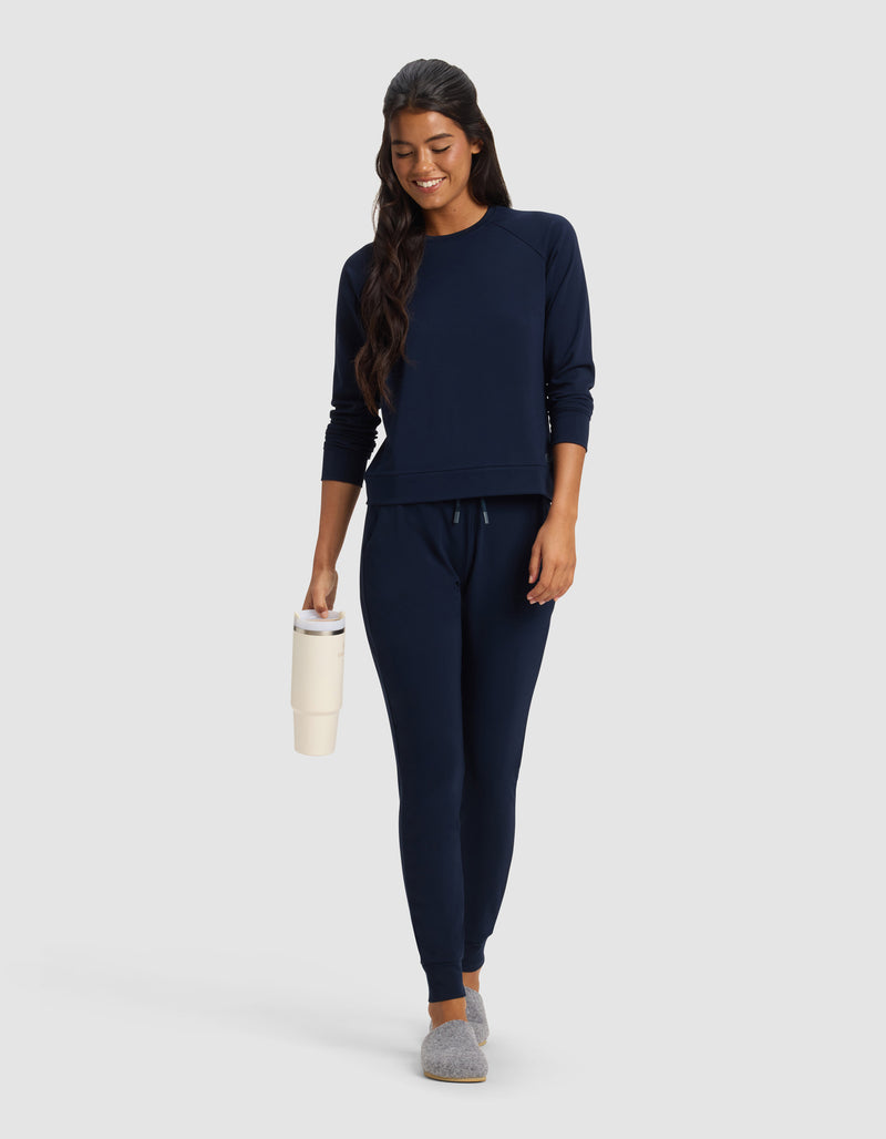 A woman with long dark hair smiles against a plain light background, wearing Cozy Earth's Women's Brushed Bamboo Jogger Pant in navy blue, paired with a matching sweatshirt, gray slippers, and holding a cream-colored tumbler.