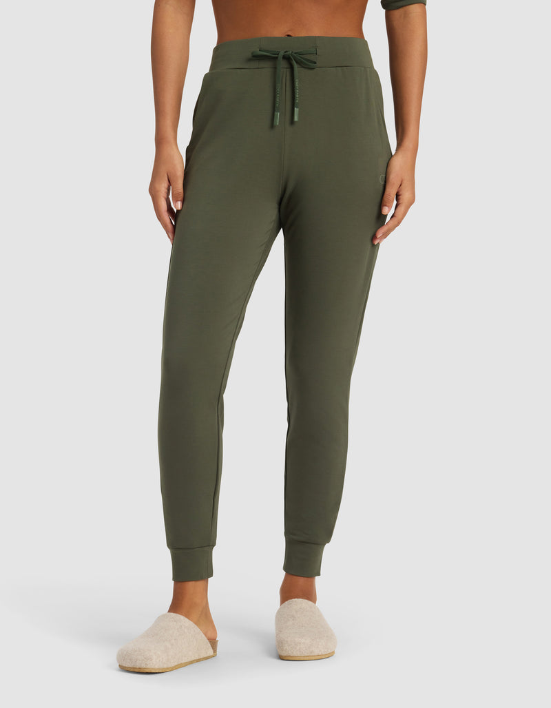 A person wears Cozy Earth's Women's Brushed Bamboo Jogger Pant in olive, featuring a drawstring waist and side pockets, paired with light beige slippers. Only the lower torso and legs are visible against a plain background.