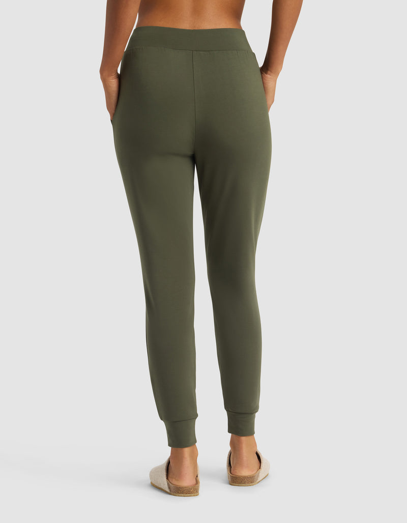 A person is seen from the back with hands in pockets, wearing Cozy Earth Women's Brushed Bamboo Jogger Pant in olive green and beige slip-on sandals against a plain white background.