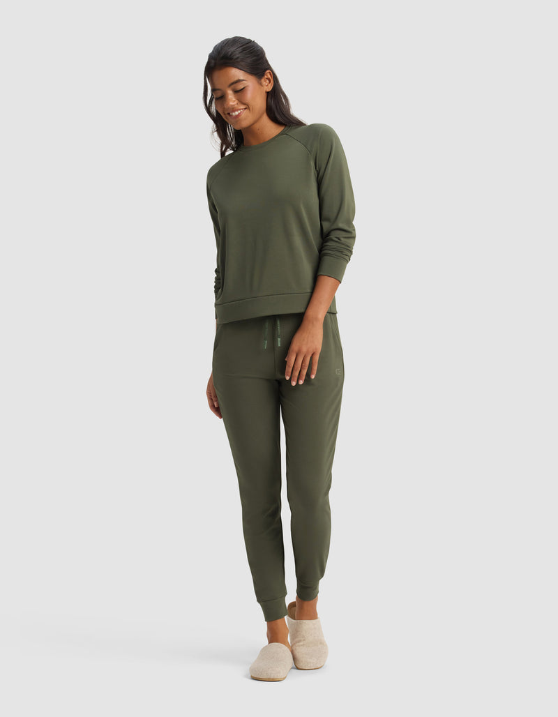 A woman smiles in Cozy Earth's Women's Brushed Bamboo Jogger Pant, paired with a matching olive green sweatshirt and beige slippers, set against a plain light background.