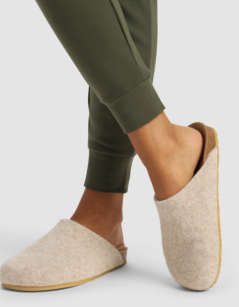A woman models the Cozy Earth Women's Brushed Bamboo Jogger Pant in olive green, paired with light beige wool slippers, standing with one foot crossed over the other against a plain background.