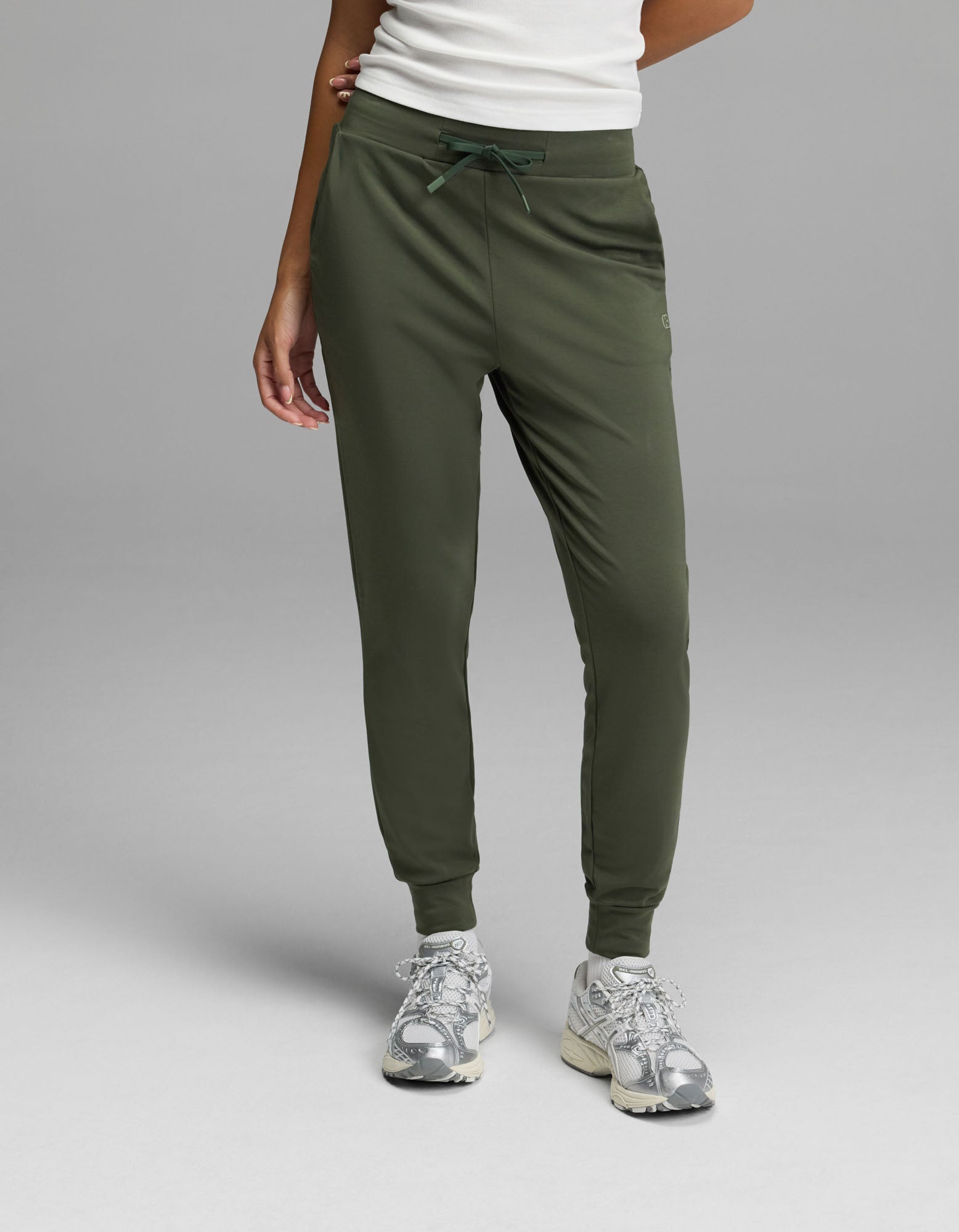 A person is shown from the waist down, wearing Cozy Earth’s Women’s Brushed Bamboo Jogger in olive green with a white top and silver sneakers, standing against a plain gray background. Only the lower body and part of one arm are visible.