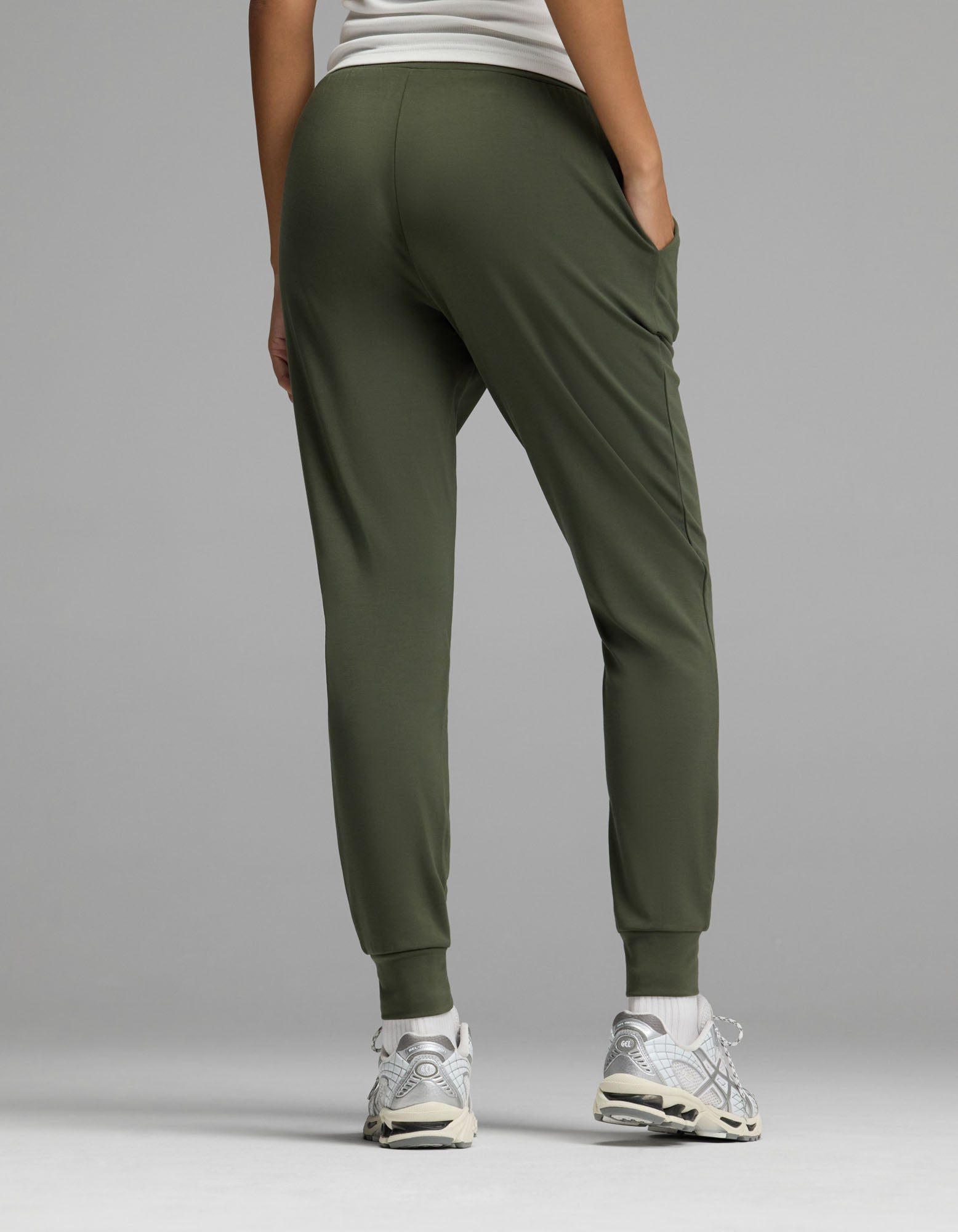 A model is shown from the back against a plain gray background, wearing Cozy Earth’s Women’s Brushed Bamboo Jogger in olive green paired with a white top and white sneakers.