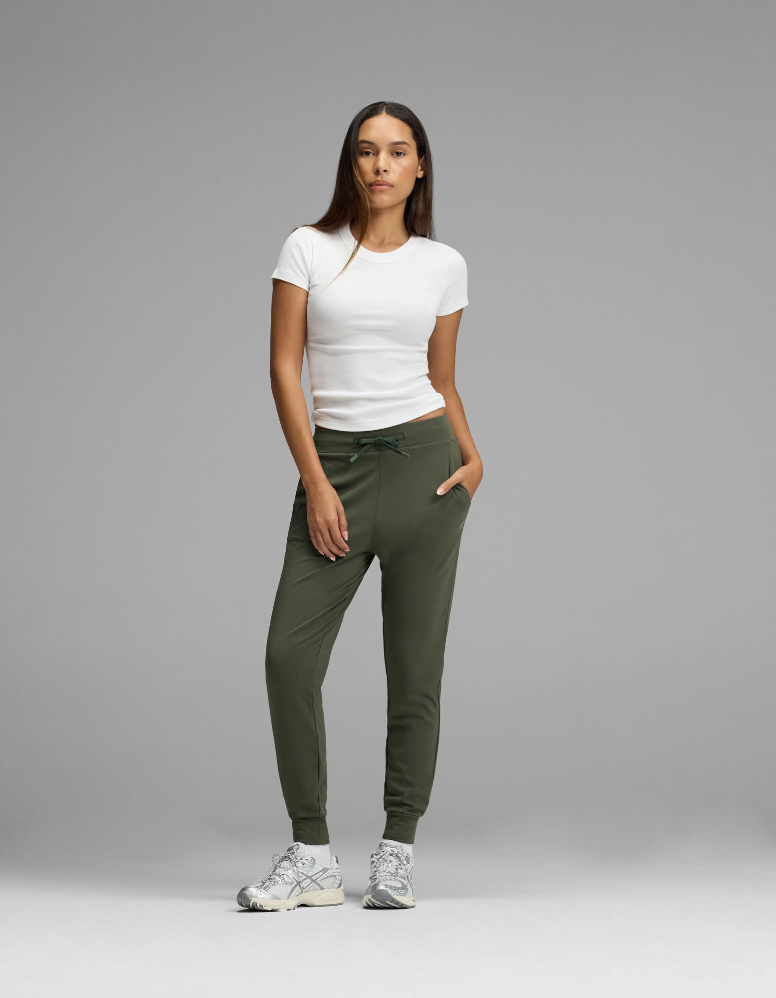 A woman stands against a gray background wearing a white t-shirt, gray sneakers, and Cozy Earth’s Women’s Brushed Bamboo Jogger in olive green, with her hands in her pockets.