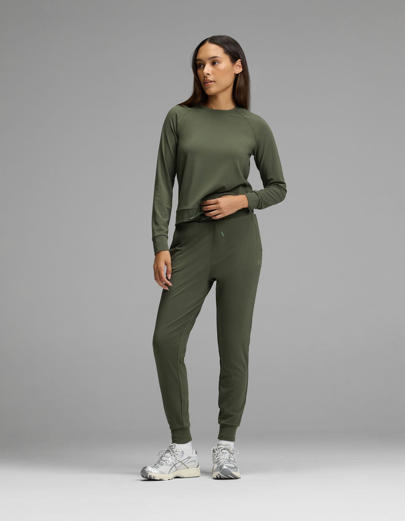 A woman stands against a plain gray background, dressed in the Cozy Earth Women’s Brushed Bamboo Jogger Set in Olive and white sneakers.|Color:Olive