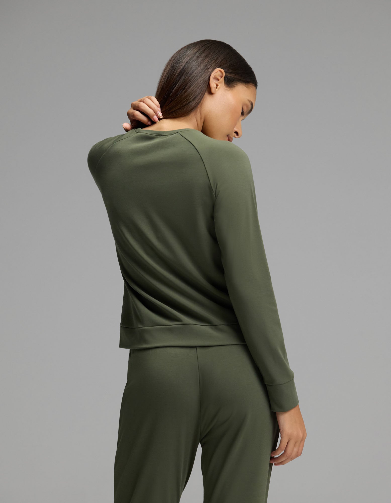 A person wearing the Cozy Earth Women’s Brushed Bamboo Jogger Set in olive green stands facing away, one hand touching the back of their neck, against a plain gray background.