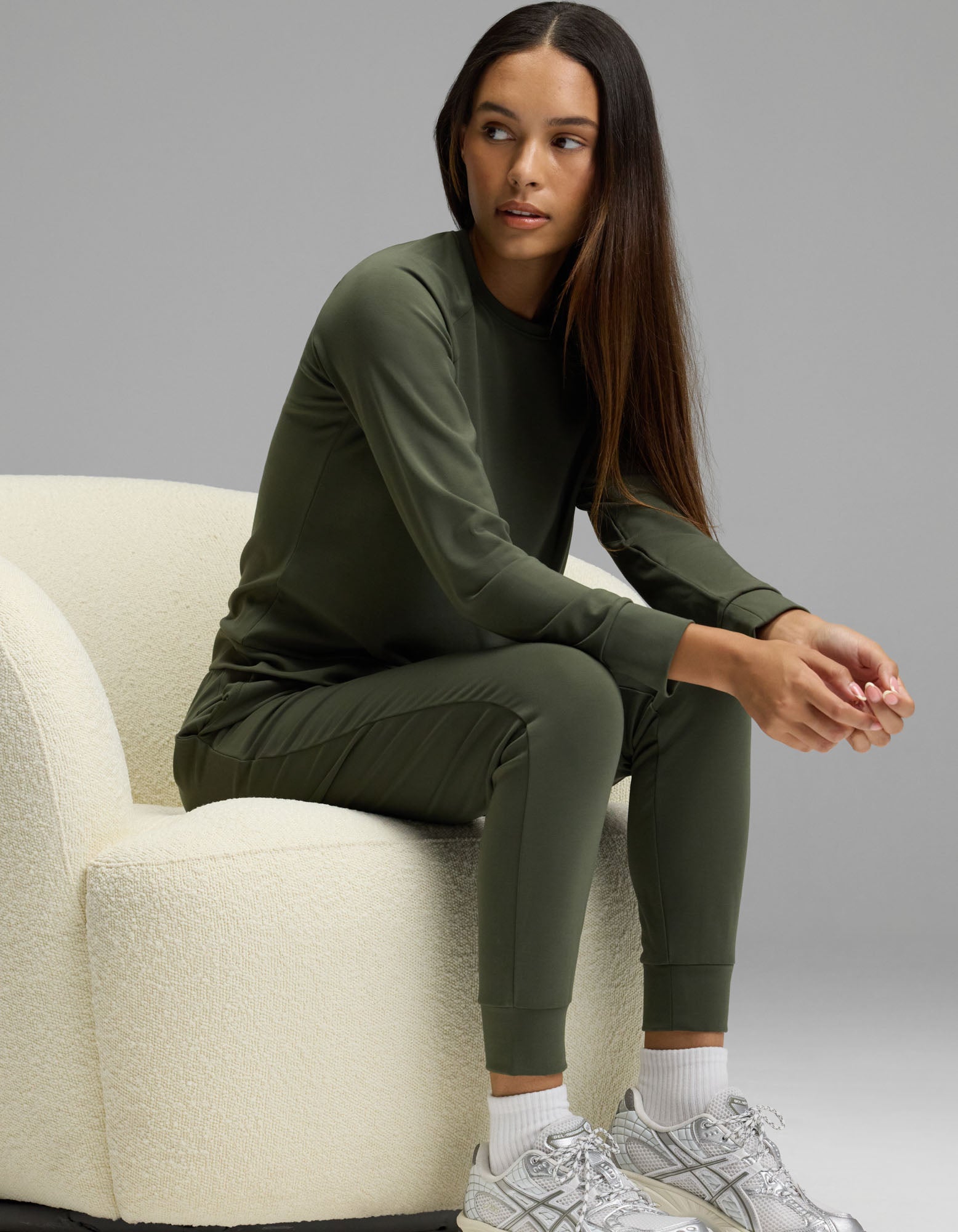 A woman wearing the Cozy Earth Women’s Brushed Bamboo Jogger Set in olive green sits on a cream-colored chair, looking to her left. She has long brown hair and pairs the outfit with white socks and silver sneakers against a plain gray background.