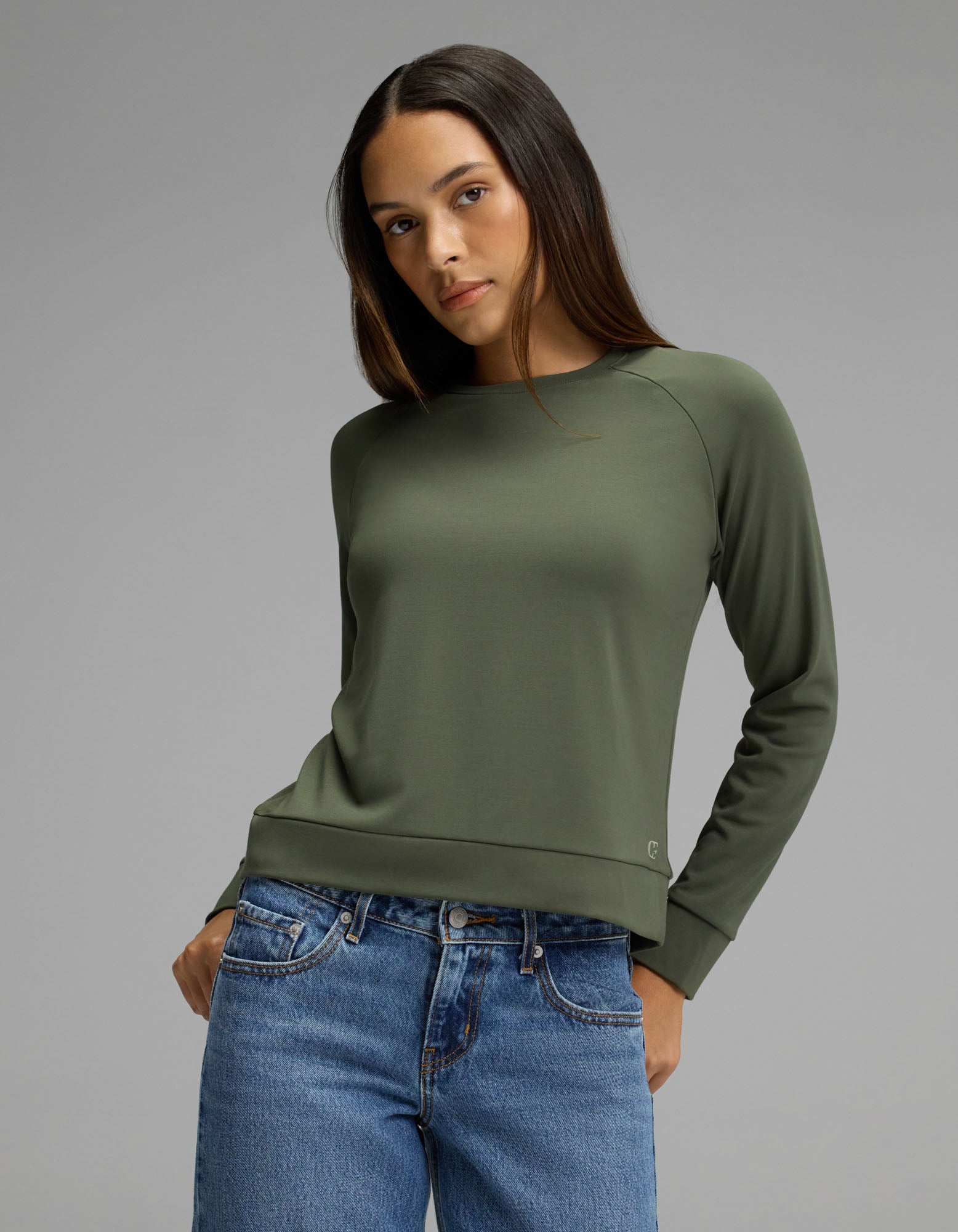 A woman models the Cozy Earth Women’s Brushed Bamboo Pullover Crew in olive, paired with blue jeans, as she stands against a plain gray background and looks slightly to the side with one hand in her pocket.