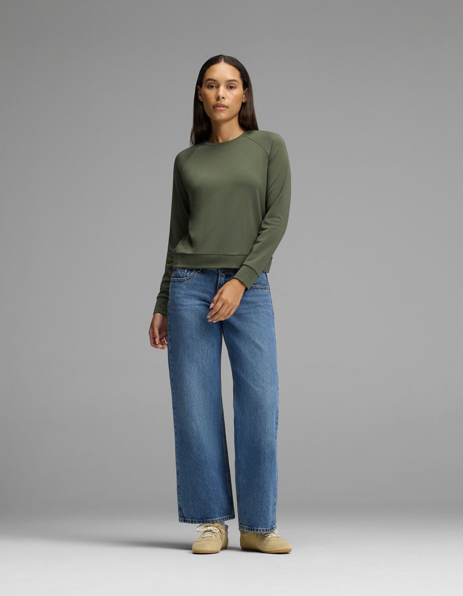 A woman stands against a gray background, wearing the Cozy Earth Women’s Brushed Bamboo Pullover Crew in olive green, paired with loose blue jeans and beige shoes. She has straight dark hair and a neutral expression, with her arms relaxed by her sides.