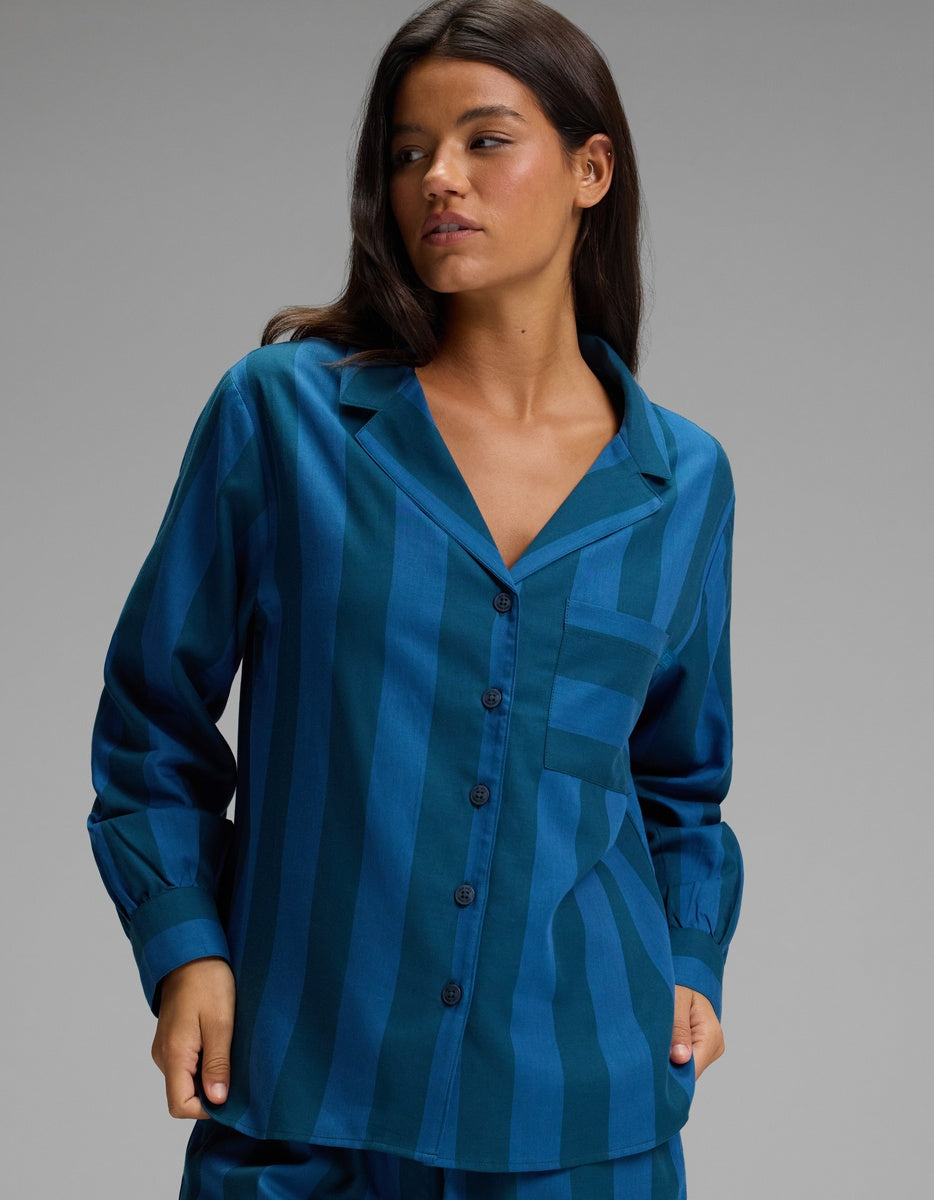 A woman wearing the Cozy Earth Women’s Belmont Long Sleeve Pajama Top in a teal and blue striped pattern stands against a plain gray background, looking to her left with one hand in her pocket.|Color:Anchor Stripe