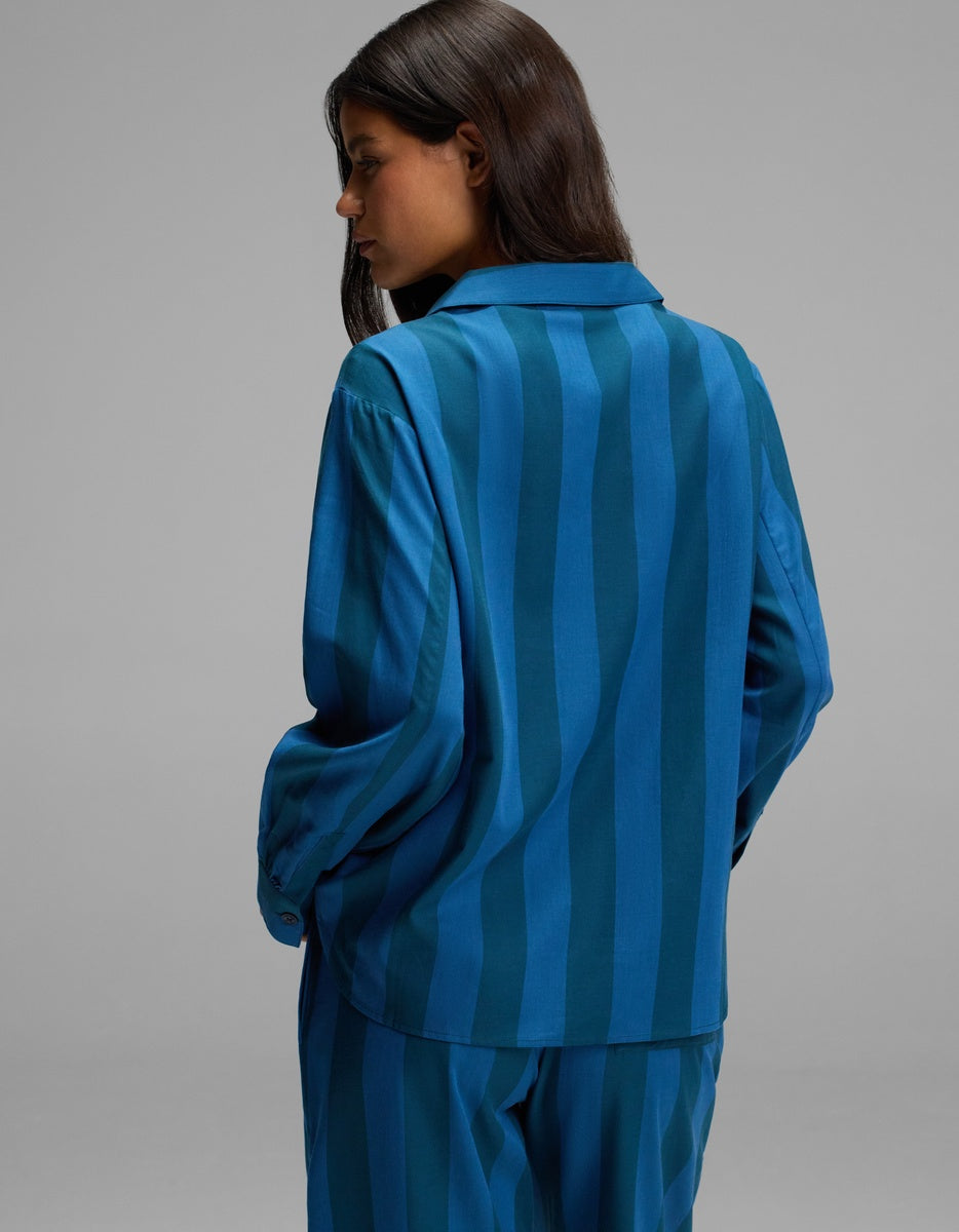 A woman with long dark hair wears the Cozy Earth Women’s Belmont Long Sleeve Pajama Top, a loose-fitting, blue striped shirt paired with matching pants, standing with her back partly turned and head slightly bowed against a plain gray background.